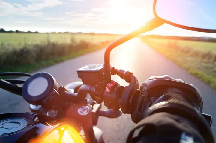 Close-up of a motorcycle handlebar with a road among green fields ahead.
